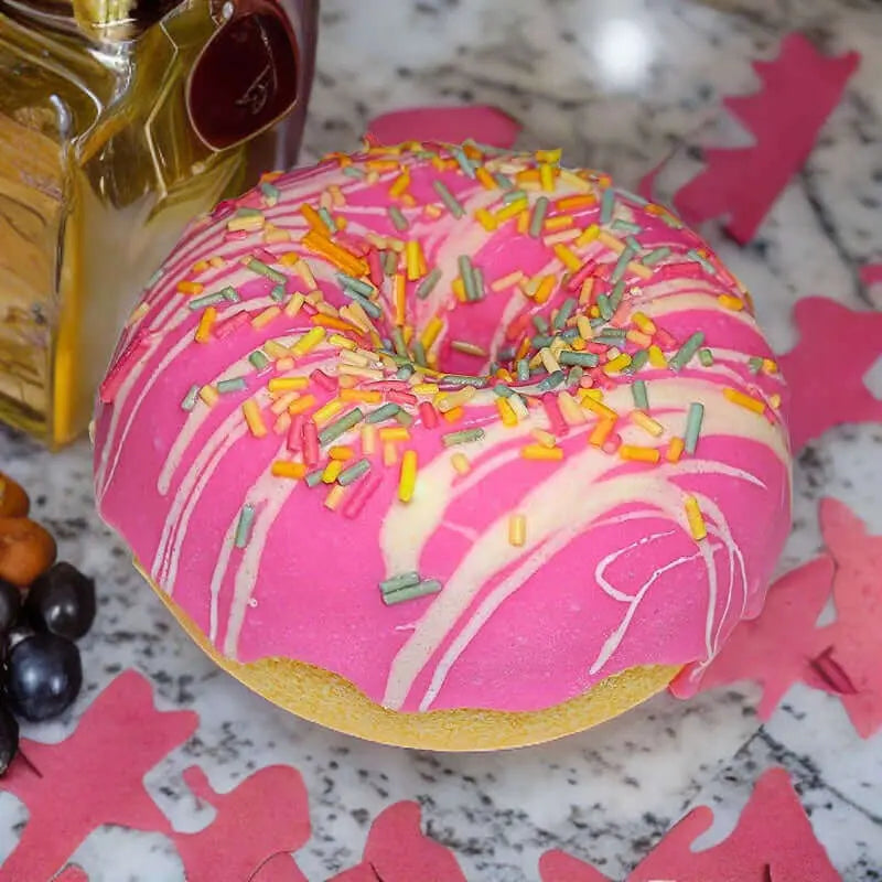 A pink-frosted donut with multicolored sprinkles and white icing drizzle sits on a marbled surface, resembling the Posh Brats Berry Vanilla Sprinkle Donut Bath Bomb, surrounded by a bottle, berries, and pink paper cutouts for a luxurious bath experience.