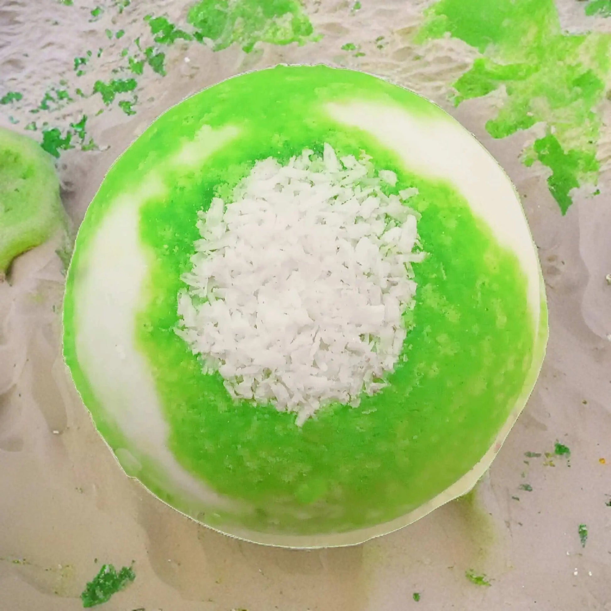 The Coconut Lime Fizzy Bath Bomb by Posh Brats features a spherical design with a bright green and white gradient, topped with white shredded coconut. It evokes a tropical paradise on a sandy surface, with green crumbs scattered around to enhance its vibrant look.