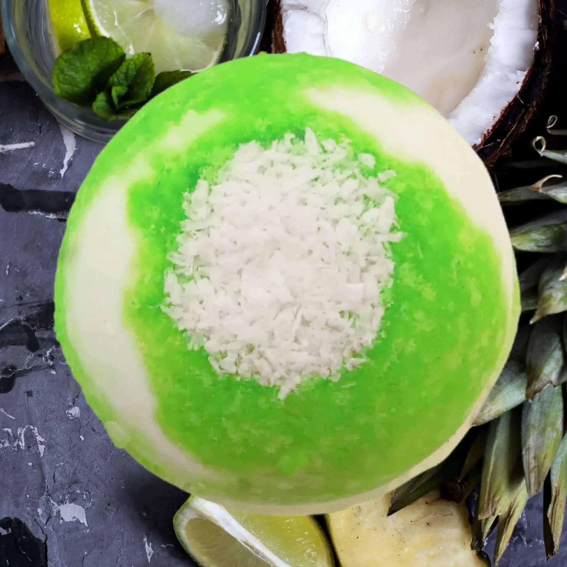 The Coconut Lime Fizzy Bath Bomb by Posh Brats evokes a tropical paradise, featuring green apple sorbet with a coconut center, surrounded by fresh mint leaves, lime slices, pineapple, and a coconut half on a textured gray surface.