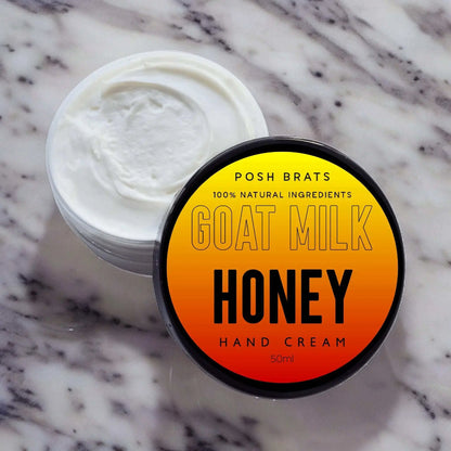 A round container of Posh Brats Goat Milk Aloe Vera Honey Soothing Hand Cream with a black lid. The yellow-to-orange gradient label hints at its soothing blend. Open on marble, it reveals a creamy mixture enriched with honey and goat milk.