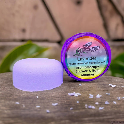 Posh Brats Lavender Aromatherapy Shower Steamer rests on a wooden surface, its colorful package adorned with lavender illustrations. The text Lavender: Pure essential oil, Aromatherapy Shower & Bath Steamer is surrounded by scattered lavender petals, evoking a calming scent.