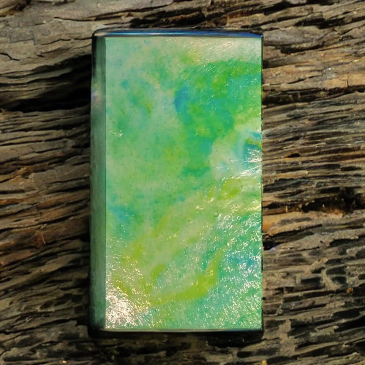 A rectangular piece of green and blue opal with a polished finish rests on textured wood, resembling the sky or sea and capturing the essence of Posh Brats Le Maquis Corsican Maquis Sugar Scrub Bars Mediterranean fragrance.