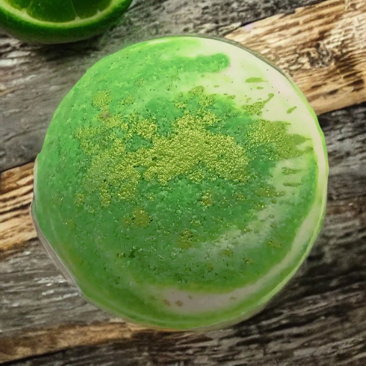 A top view of a creamy, swirled drink adorned with gold powder on a rustic wooden surface beside a half-visible lime evokes the zesty vibrance of Posh Brats Frozen Lime Margarita Fizzy Bath Bomb.