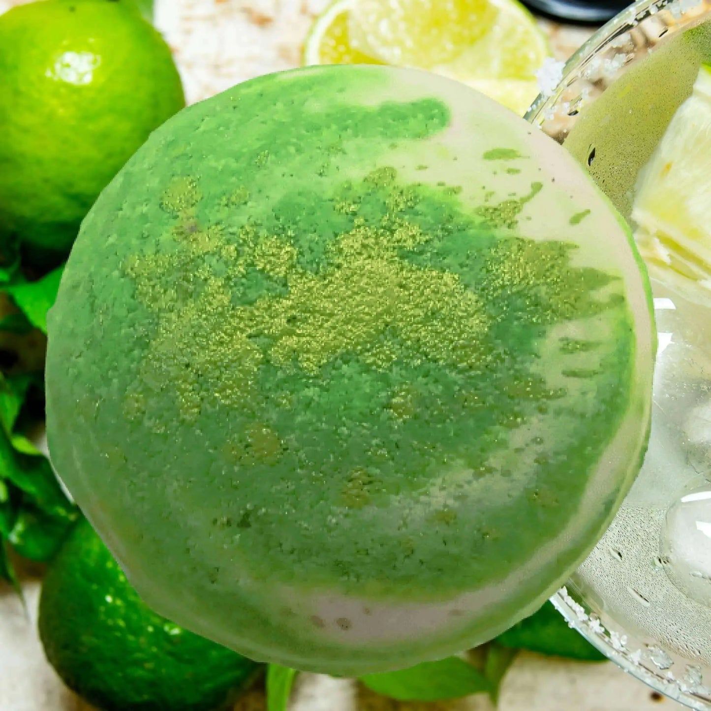 A lush green margarita glass, rimmed with salt and filled with zesty tequila, is surrounded by fresh limes. Overhead, the gradient of green hues reflects the tangy vibrance of the Posh Brats Frozen Lime Margarita Fizzy Bath Bomb in the background.