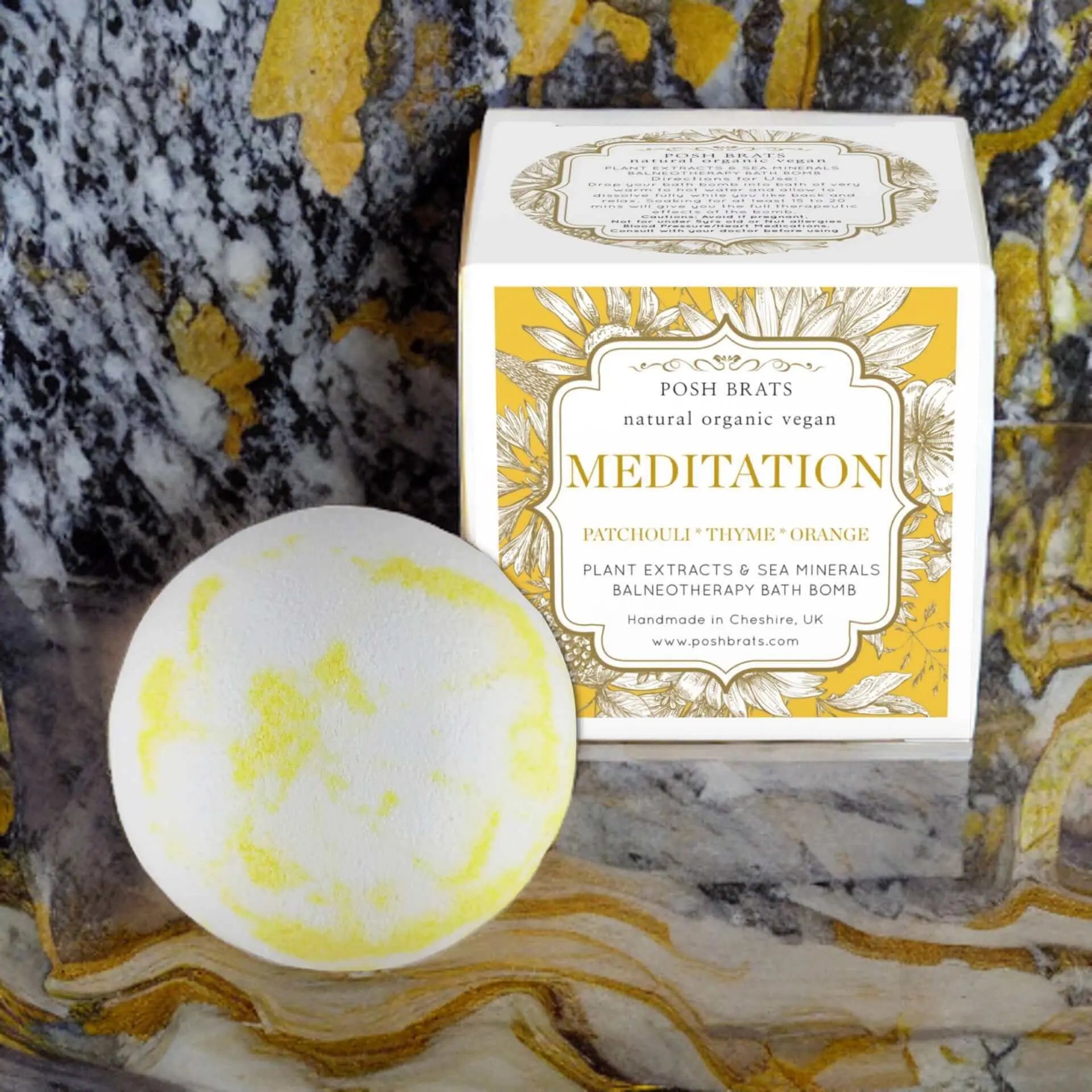 A round bath bomb with yellow accents by Posh Brats rests beside a Meditation Aromatherapy Bath Bomb box. It boasts patchouli, thyme, and orange essential oils with plant extracts and sea minerals for relaxation on the marbled stone surface.