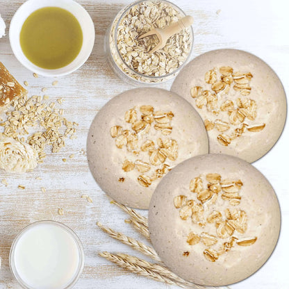 Three round oatmeal cookies rest on a light wooden surface alongside Posh Brats Skin Soothing Milk Oats Unscented Fizzy Bath Bomb, scattered oats, wheat stalks, a small bowl of olive oil, and a glass of milk.