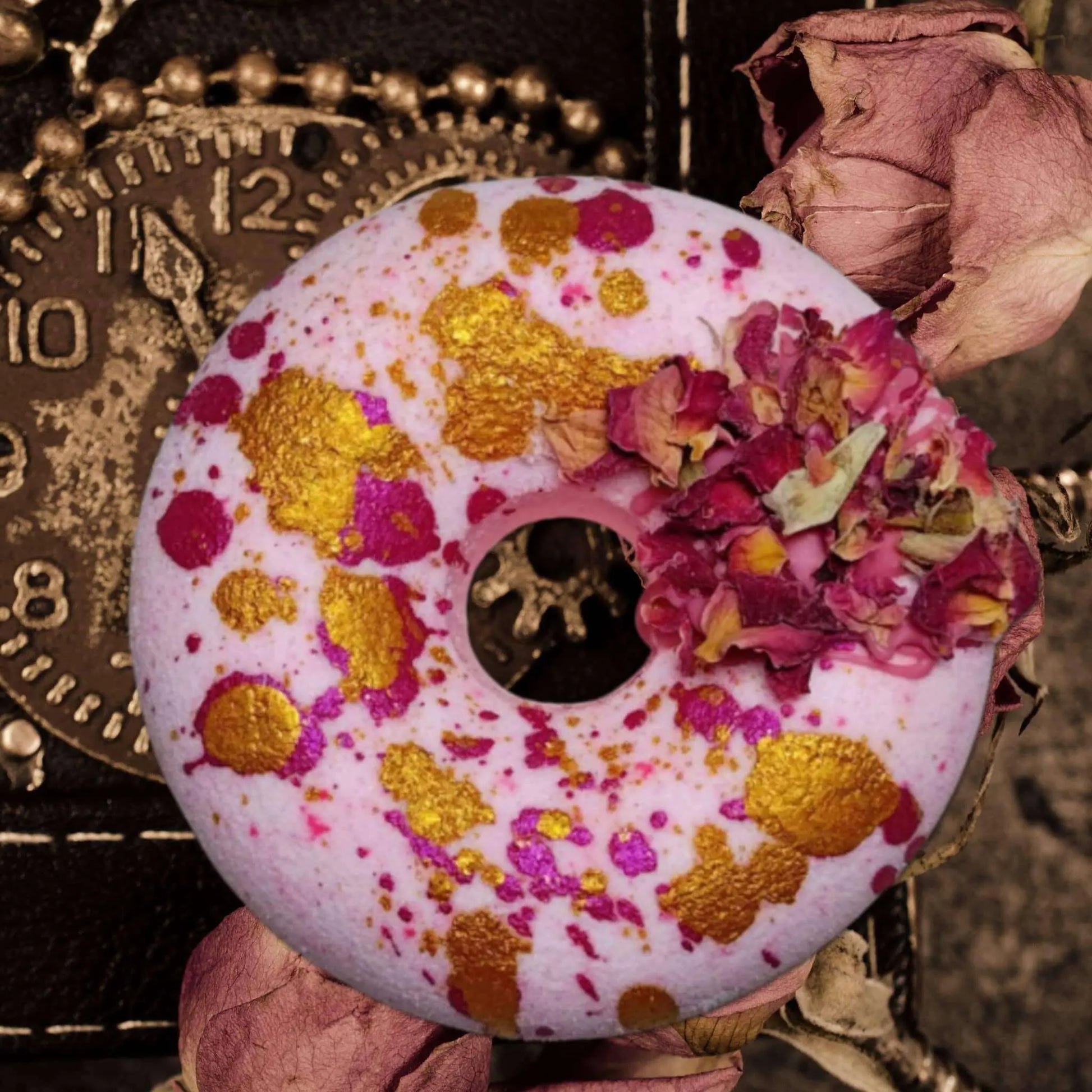 The Moonlight Kiss Donut Bath Bomb by Posh Brats is a round, pink bomb with gold and magenta splatters, adorned with dried flower petals. It sits on a vintage clock face amidst dried pink roses, creating an aromatic journey.