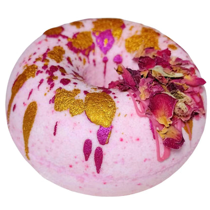 Introducing the Moonlight Kiss Donut Bath Bomb by Posh Brats: a pink doughnut-shaped delight accented with gold splashes and dried rose petals, featuring vibrant magenta drips. Indulge in a luxurious aromatic journey.