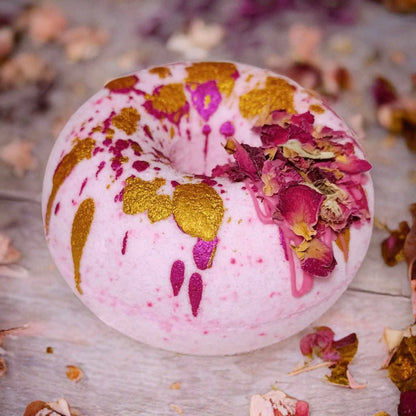 A round, pink Posh Brats Moonlight Kiss Donut Bath Bomb with gold splatters and dried red rose petals sits on a light wooden surface, an aromatic experience amidst scattered rose petals.