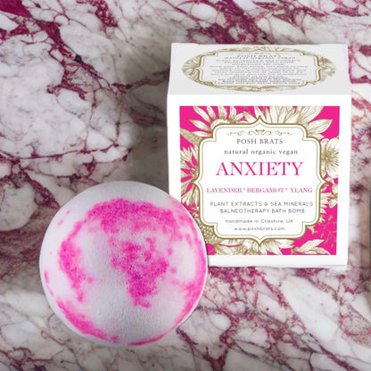 A round pink and white Posh Brats Anxiety Aromatherapy Bath Bomb sits next to a stylish white box with gold and pink accents. Perfect for anxiety relief, its set against a marbled backdrop for ultimate relaxation.