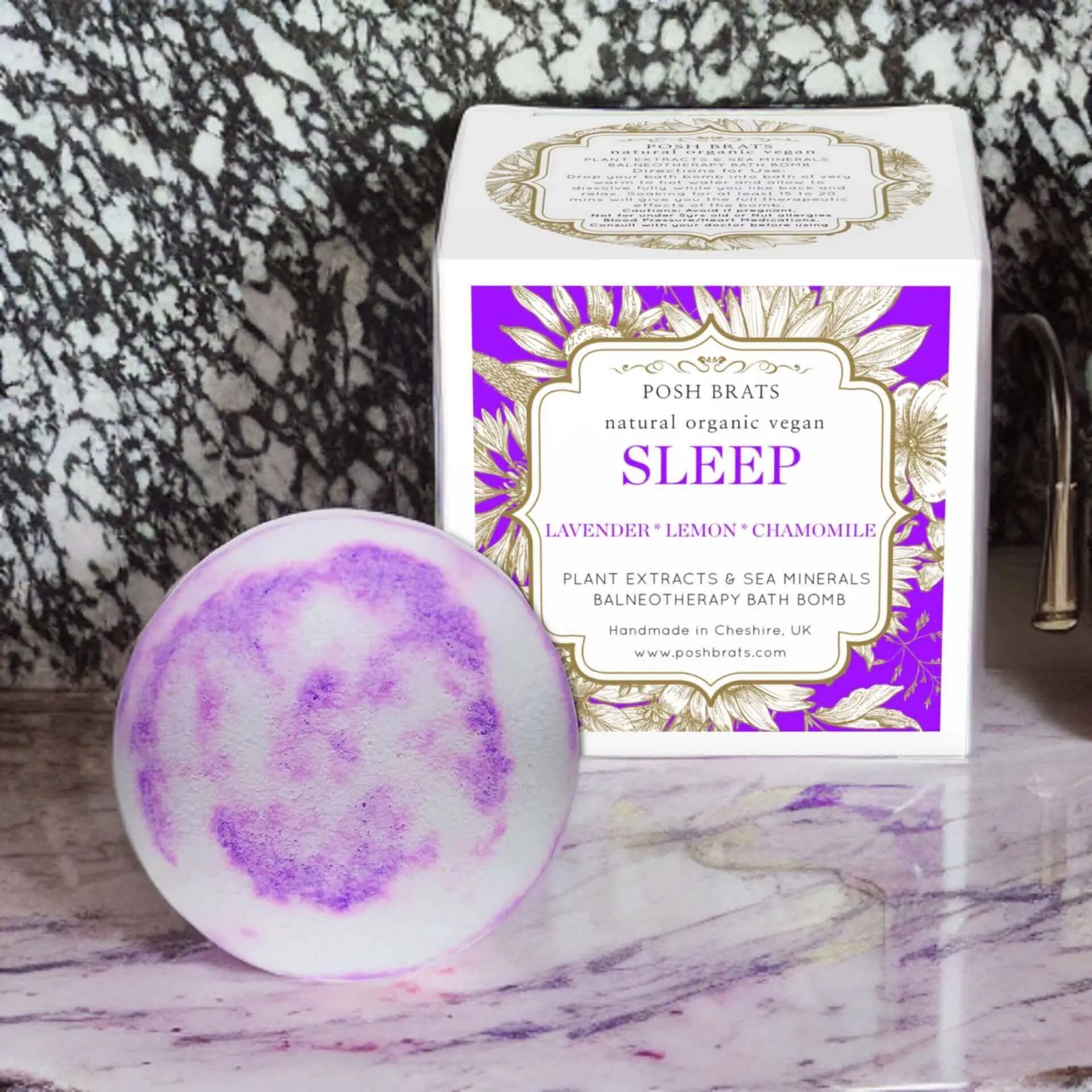 A Sleep Aromatherapy Bath Bomb by Posh Brats, with purple and white swirls, sits next to a Sleep box infused with lavender, lemon, and chamomile essential oils. The packaging features gold floral designs, all elegantly placed on a sleek marble surface for the ultimate relaxation experience.